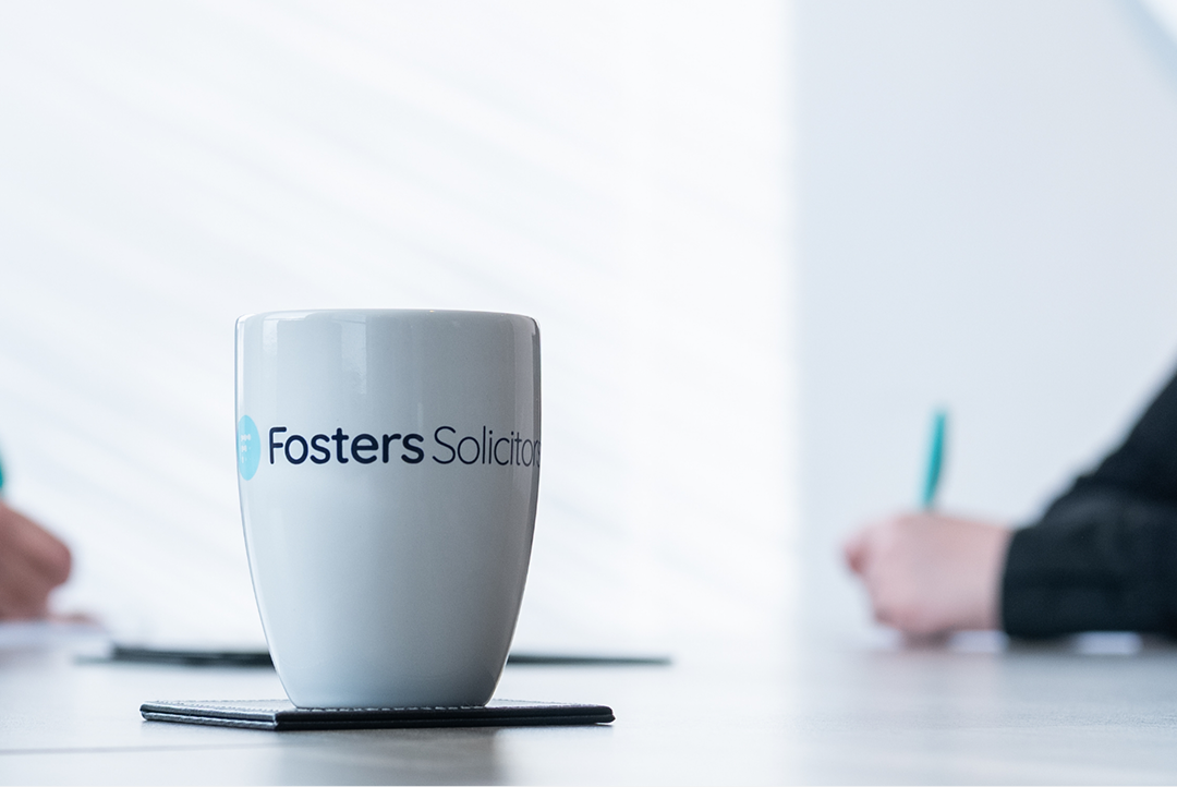 Fosters Solicitors mug on a table with a colleague writing notes in a bright office
