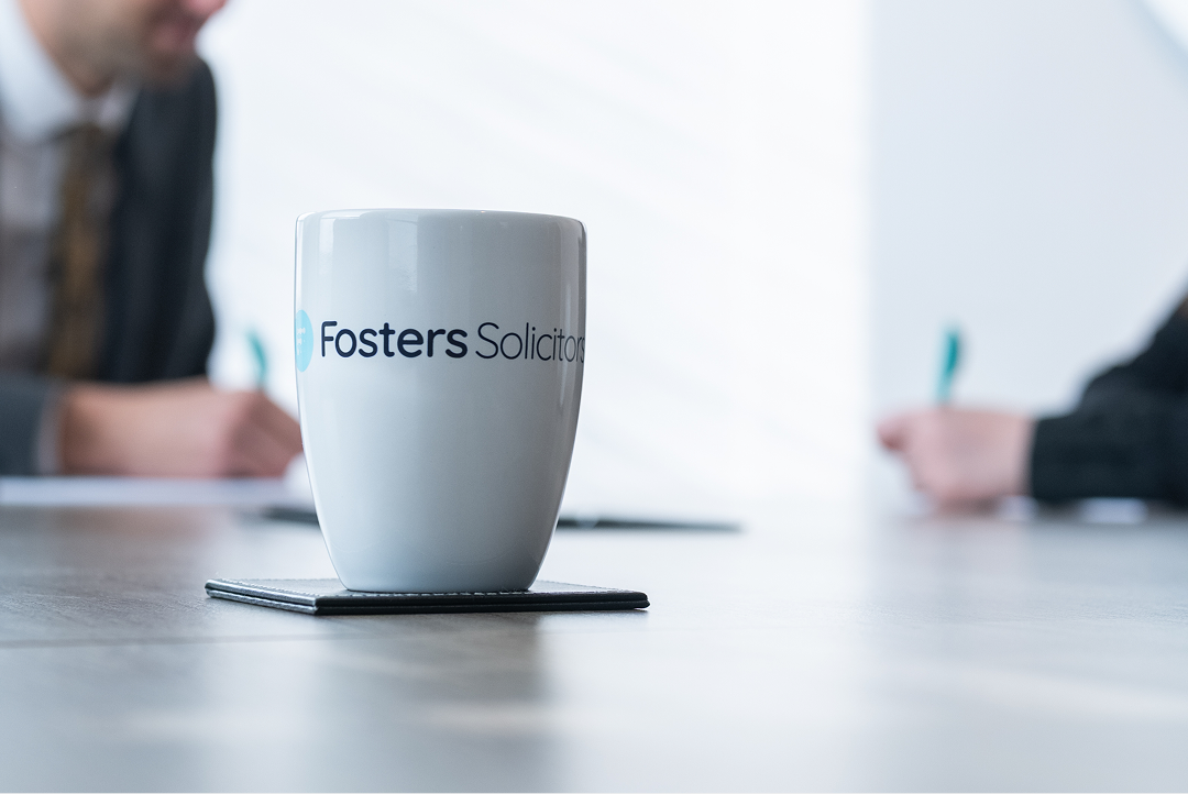 Fosters Solicitors mug on a table with two colleagues in the background