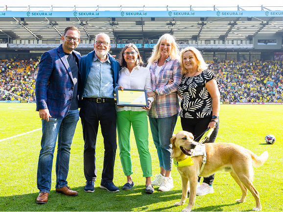 Fosters Solicitors representatives with a guide dog receiving a community award pitchside at Carrow Road