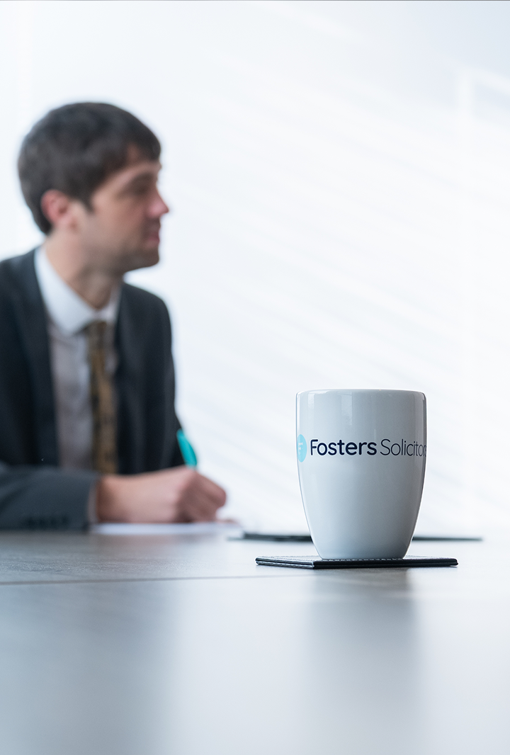 Fosters Solicitors mug on a table with a solicitor writing notes in the background